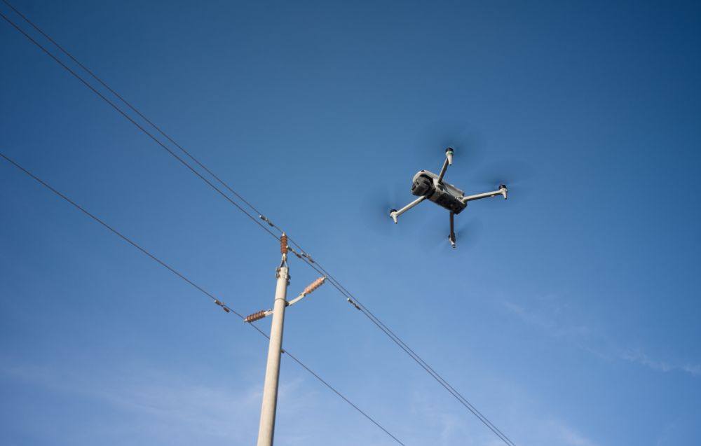 Dron przemysłowy DJI w locie obok linii energetycznych na tle niebieskiego nieba.